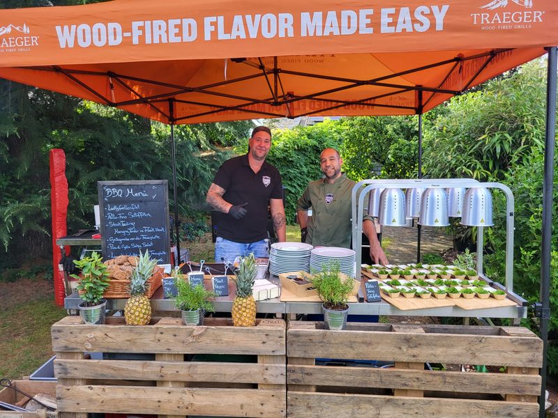 Zwei Mitarbeiter von Hybrid BBQ hinter dem Buffet mit BBQ-Menü-Tafel unter dem orangen Zelt