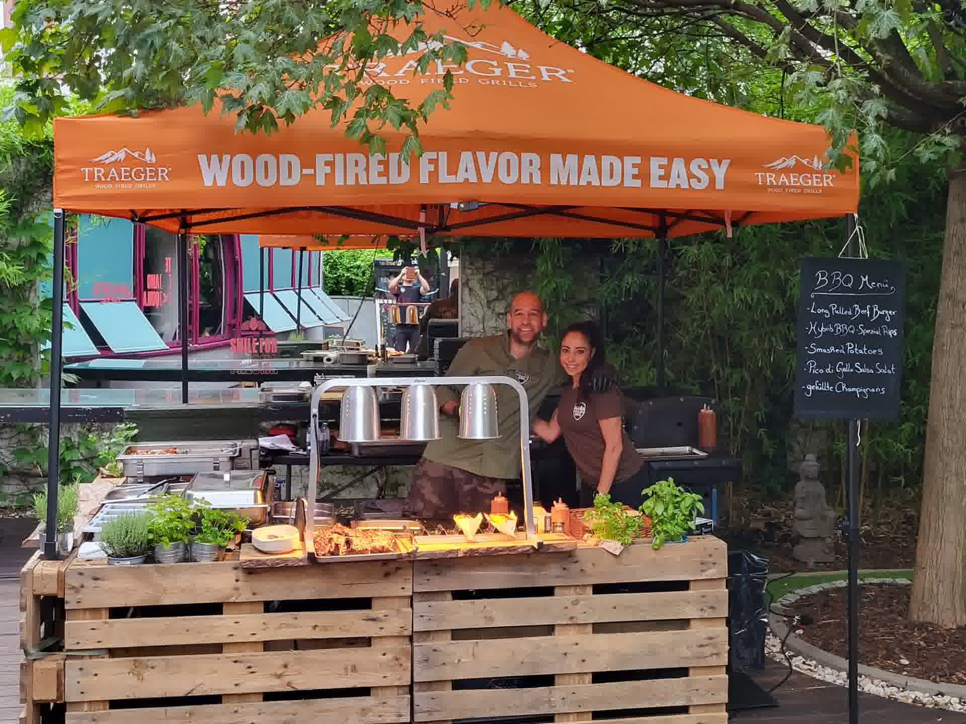Der Hybrid-BBQ-Events-Stand unter orangem Traeger-Zelt mit Team und Grillbuffet auf einem Event