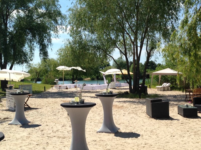 Sandstrand am See mit weiß gedeckter Tafel, Stehtischen und Sonnenschirmen für eine Hochzeitsfeier